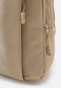 Beige ryggsäck gjord av texturerad tyg, med en dragkedjeficka och dragband. Slät yta med subtila sömnadsdetaljer.