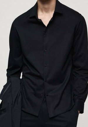 Homme portant une chemise noire à manches longues avec col et boutons, les mains dans les poches, tenant une veste noire sur un bras.