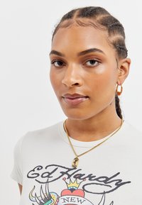 Lys grå t-skjorte med svart grafisk trykk, gullkjede med anheng, gull hoops øredobber. Håret stylet i flettede seksjoner.