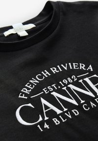 Schwarzes T-Shirt mit Rundhalsausschnitt und weißem Stickschriftzug: „French Riviera Est. 1982 CANNES 14 BLVD CARL“ auf der Vorderseite.