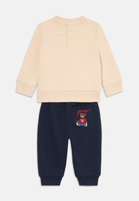 Beige langermet topp med knappelukking; marineblå joggebukser med bjørnebroderi og rødt "guess"-tekst på baklommen. Mykt bomullsmateriale.