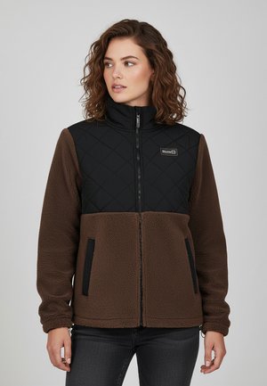 Frau mit lockigem Haar, die eine braune Fleecejacke mit schwarzem gesteppter Oberteil, Reißverschluss vorne und Seitentaschen trägt, schaut seitlich.