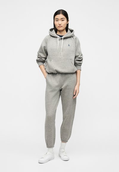 Νεαρή γυναίκα που στέκεται, φοράει γκρι μπλούζα με κουκούλα και ταιριαστό παντελόνι φόρμας, λευκές κάλτσες και αθλητικά παπούτσια, σε φόντο απλό λευκό.