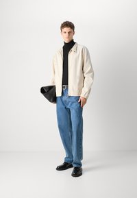 Beige Jacke, schwarzer Rollkragenpullover, blaue, lässig geschnittene Jeans und schwarze Schuhe. Modell hält eine schwarze Tasche; minimalistisches Design und lässiger Stil.