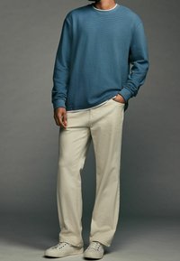Chemise bleue à manches longues avec texture côtelée, superposée sur une chemise blanche, associée à un pantalon large couleur crème et des baskets blanches.