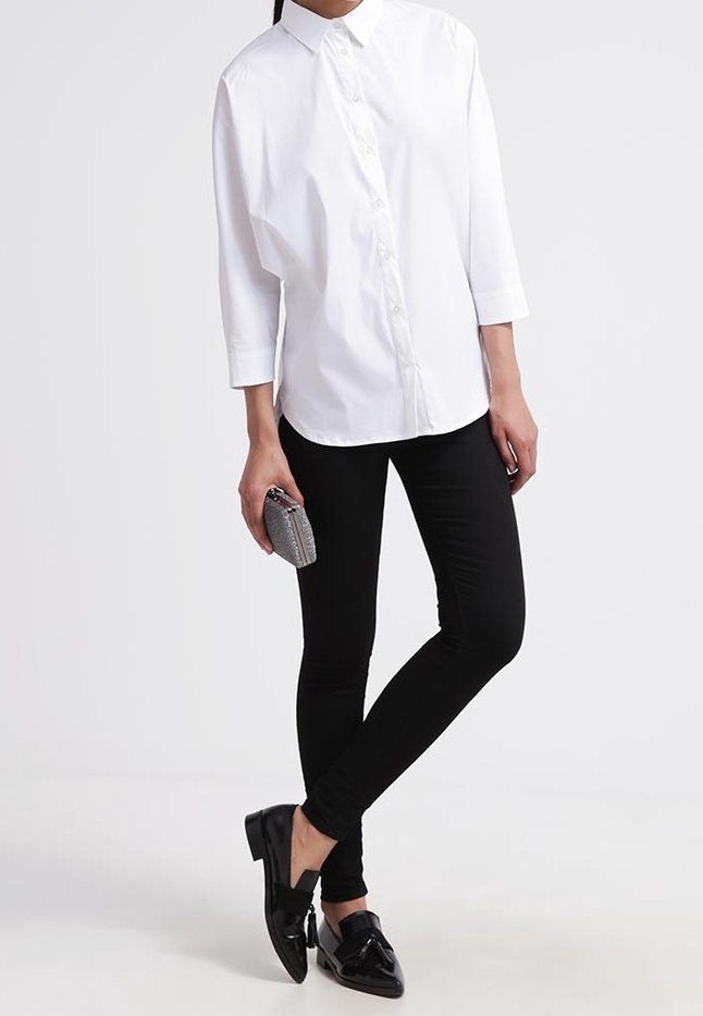 Femme portant une chemise blanche boutonnée, pantalon skinny noir, mocassins noirs, tenant une petite pochette texturée.
