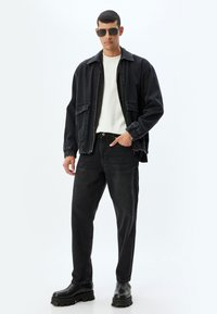 Chaqueta de mezclilla negra con dos bolsillos, camiseta blanca, vaqueros negros y botas gruesas negras. Modelo con gafas de sol de gran tamaño, posando frente a un fondo blanco.
