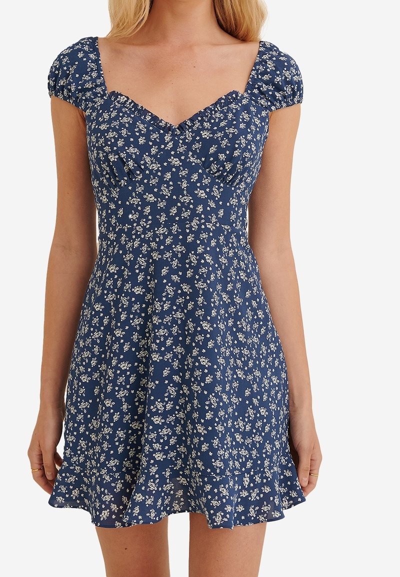 Robe courte bleue à motif floral avec manches bouffantes, corsage ajusté et encolure en cœur à volants portée par une femme des épaules jusqu'à la mi-cuisse.