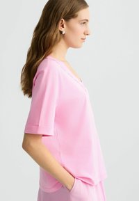 Maglietta rosa a maniche corte con scollo a V, texture morbida e orli arrotolati. Presenta un sottile dettaglio del logo lungo il collo.