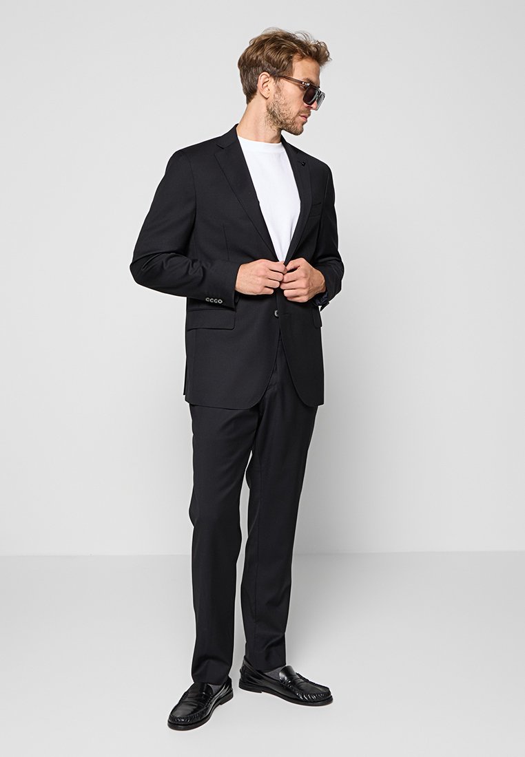 Man in een zwart kostuumjack over een wit shirt, met zwarte broek, zwarte schoenen en een zonnebril, staand tegen een effen achtergrond.