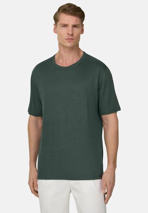Homme portant un t-shirt à manches courtes col ras du cou vert foncé et un pantalon blanc, debout devant un fond gris clair.