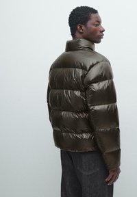 Dunkelbraune Steppjacke mit hohem Kragen und gesteppter Optik, mit glänzendem Finish und lockerem Sitz. Kombiniert mit dunkelblauer Jeanshose.