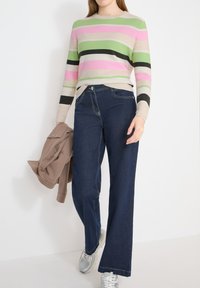 Personne portant un pull rayé rose, vert et beige avec un jean bleu foncé et des baskets blanches, tenant une veste marron dans la main gauche.