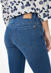 Blauwe denim jeans met een gladde textuur, vijfzakkenontwerp en een leren merklabel op de achterkant van de tailleband. Close-up van de achterkant.