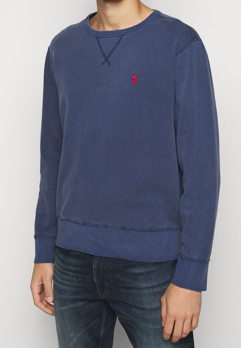 Man i en bleknad marinblå långärmad sweatshirt med en liten röd broderad logotyp på bröstet, ihop med mörkblå jeans.