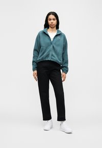 Γυναίκα που στέκεται, φοράει γαλάζιο φλις μπουφάν, λευκό μπλουζάκι, μαύρο παντελόνι και λευκά αθλητικά παπούτσια, με φόντο απλό και φωτεινό.