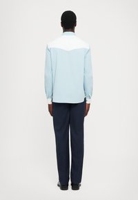 Camicia azzurro chiaro con yoke bianco, abbinata a pantaloni navy scuri. Taglio slim, maniche lunghe con bottoni ai polsini, tessuto dalla texture liscia.