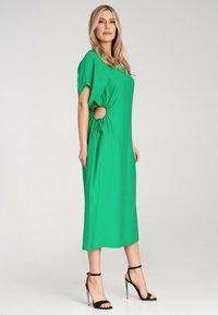 Grünes Midi-Kleid mit kurzen Ärmeln, mit Seitenausschnitten und einer Kordelzugdetail in der Taille. Glattes Material und gerade Silhouette.
