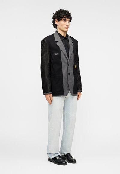 Σακάκι με αντίθετες μαύρες στενές ρίγες και γκρι ύφασμα, μονόκουμπο με τρία κουμπιά, σετάρισμένο με ανοιχτόχρωμο τζιν και μαύρα παπούτσια.