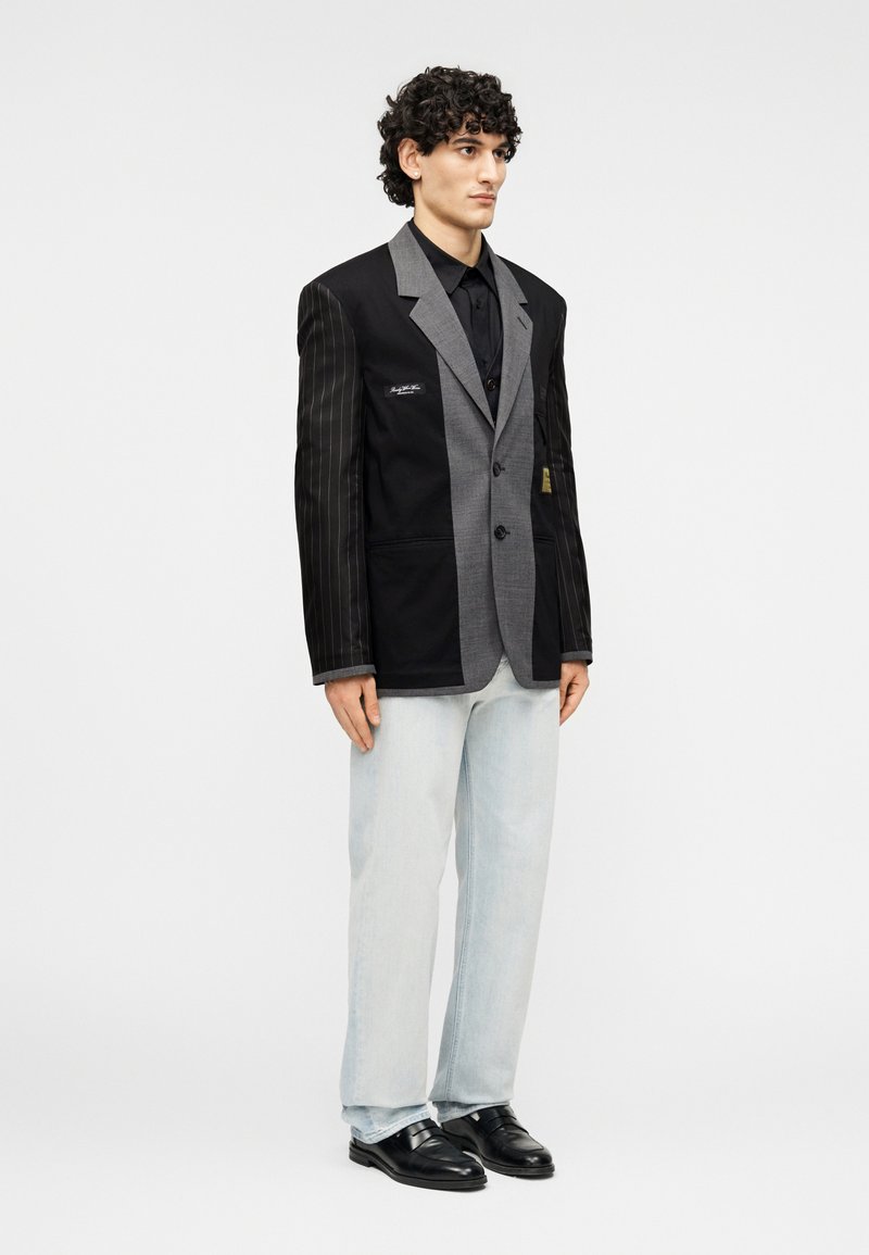 Blazer en tissu gris avec fines rayures noires contrastantes, à simple boutonnage avec trois boutons, porté avec un jean clair et des chaussures noires.