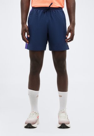 Mężczyzna noszący granatowe sportowe spodenki, białe skarpetki za kostkę oraz białe buty do biegania, stojący na neutralnym tle.