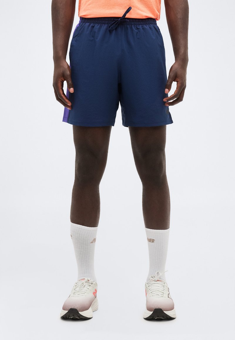 Άνδρας που φοράει σκούρο μπλε αθλητικό σορτς, λευκές κάλτσες πάνω από τον αστράγαλο και λευκά παπούτσια για τρέξιμο, στέκεται μπροστά σε ουδέτερο φόντο.