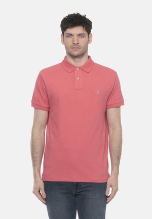 Uomo che indossa una polo a maniche corte color salmone con colletto e bottoni, in piedi dritto contro uno sfondo bianco uniforme.