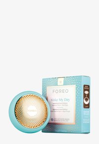 FOREO UFO-enhet i teal med en gyllene cirkelår topp, visad bredvid en ljusblå och vit låda märkt "Make My Day" med gyllene accenter.