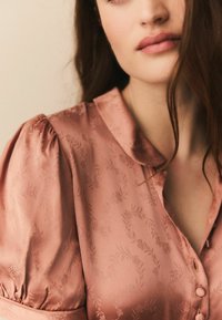 Blush satin bluse med krave, pufærmer og subtilt blomstermønster; har knaplukning foran og blød, glat tekstur.
