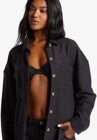 Sort skjorte med knapærmer lavet af tekstureret stof. Har en stor lomme på forsiden og runde knapper. Modellen bærer en sort bikini under.