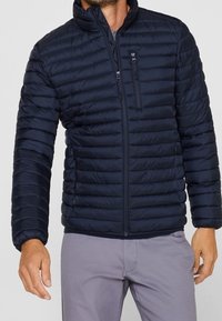 Mørkeblå puff jakke med vandret syninger, høj krave, frontlynlås og en lynlåslomme på brystet. Letvægts og quiltede design.