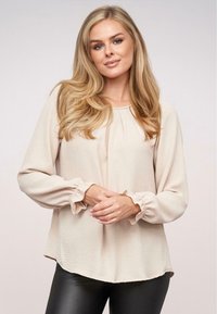 Blusa beige a maniche lunghe con dettagli in cuffia arricciata, colletto rotondo e tessuto leggermente testurizzato, abbinata a pantaloni neri aderenti.