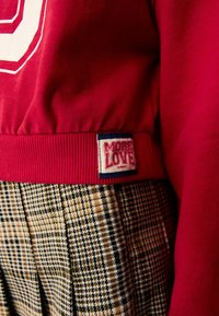 Rød sweatshirt med ribbet kant og et "More Love" patch. Kombination med plisserede bukser i beige og sort houndstooth mønster.