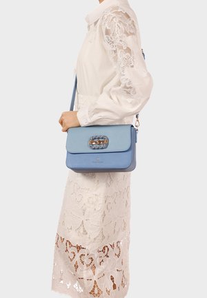 Donna con un vestito di pizzo bianco che indossa una borsa a tracolla azzurro chiaro con una chiusura a catena d'oro all'altezza della vita.
