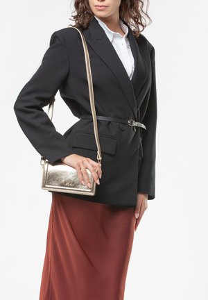 Mujer con blazer negro ceñido con cinturón y falda de color óxido, llevando un bolso cruzado metálico dorado sobre el hombro.