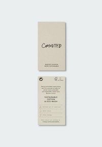 Deux étiquettes de vêtements beige avec un texte sur la durabilité, mettant en avant le coton durable, le lavage écologique, la réduction des produits chimiques, les économies d'eau et d'énergie.
