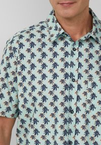 Korte mouwen overhemd met knopen in lichtblauw met een patroon van marineblauwe palmbomen en oranje accenten, voorzien van een kleine voorkantzak en zwarte knopen.
