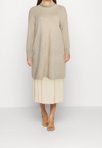 Krémová, dlouhá pletená tunika s vysokým límcem, spárovaná s plisovanou, světlou sukní a sandály s pásky v béžové barvě.