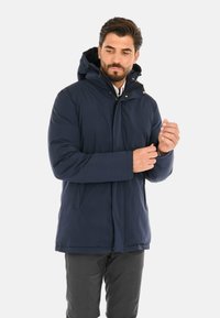 Giacca impermeabile blu navy con cappuccio, dotata di una chiusura a zip frontale, dettagli con bottoni a pressione e due tasche laterali. Texture liscia, design lunghezza ginocchio.