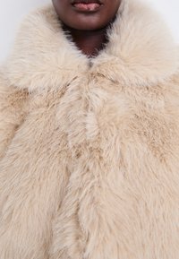 Manteau en fausse fourrure beige avec un large col, présentant une texture douce et duveteuse et une fermeture frontale simple. Aucun fermoir ou motif visible.
