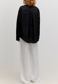 Schwarzes, oversized Hemd mit Kragen und langen Ärmeln, kombiniert mit locker sitzenden weißen Hosen, von hinten vor einem schlichten Hintergrund gezeigt.