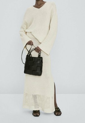 Personne portant un pull en tricot crème et une longue jupe, tenant un sac à main noir tissé, avec des sandales noires visibles.