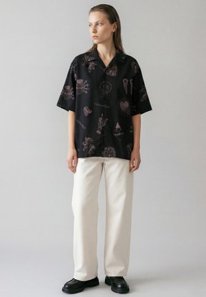 Giovane con capelli bagnati pettinati all'indietro, indossa una maglietta nera a maniche corte con disegni rosa, pantaloni bianchi e scarpe nere, in piedi in posizione neutra.