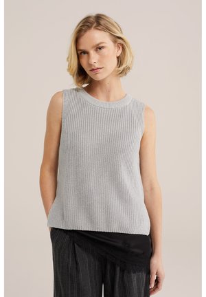Blondynka ubrana w bezrękawnik jasnoszary sweter z dzianiny oraz czarne spodnie w prążki z asymetrycznym czarnym koronkowym detalem.