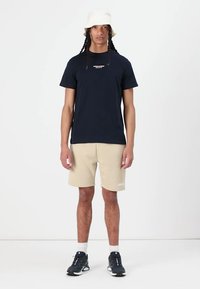 Marine T-skjorte med hvit tekst, beige shorts og svarte joggesko. Inkluderer en bøttehatt og sokker, satt mot en hvit bakgrunn.