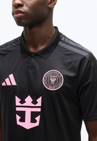 Άνδρας που φοράει μαύρη φανέλα ποδοσφαίρου με ροζ έμβλημα του Miami Club Internacional de Futbol, ροζ λογότυπο Adidas και ροζ έμβλημα κορώνας.