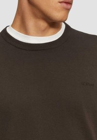 Brauner Strickpullover mit Rundhalsausschnitt und geripptem Kragen, über einem weißen Hemd getragen. Verfügt über ein dezentes Logo auf der Brust.