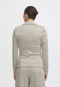 Blazer beige à carreaux avec manches longues, présentant une coupe ajustée et des revers à encoche. Tissu lisse avec une texture subtile et une coupe sur mesure.
