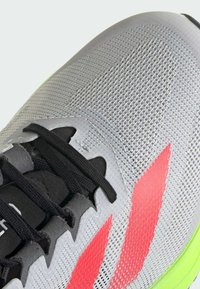 Leichte graue Sportschuh mit einem atmungsaktiven Mesh-Obermaterial, schwarzen Schnürsenkeln und neongrünen Akzenten. Mit roten Designstreifen und strukturiertem Sohlenprofil.
