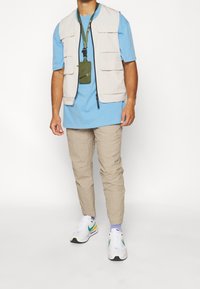 Beige väst med flera fickor över en ljusblå t-shirt, kombinerat med beige byxor med textur och vita sneakers med gröna och gula detaljer.